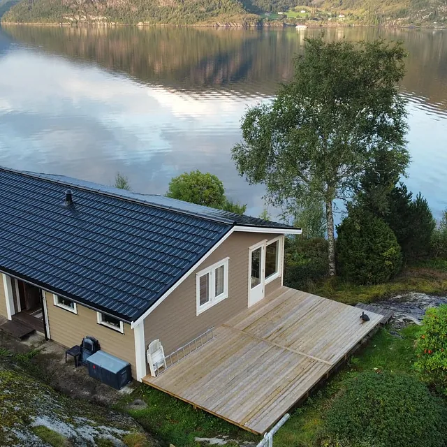Enebolig ved sjøen med nytt mørkt tak og stor treterrasse, fotografert fra luften i rolige omgivelser.