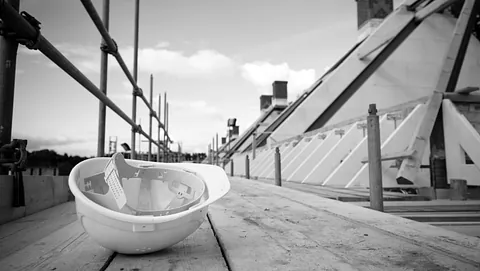Vernehjelm på stillas ved takarbeid, fotografert i sort-hvitt under byggeprosjekt.
