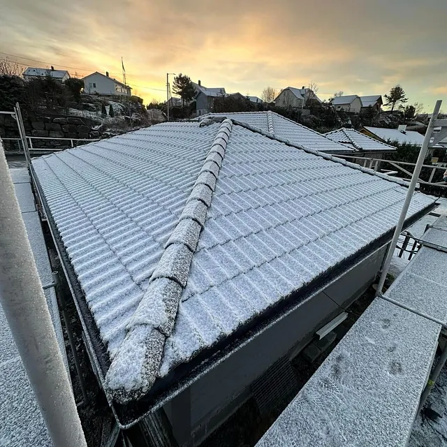 Tak med lett snødekke og møner på mørk bolig, fotografert fra stillas i vinterlys ved soloppgang.