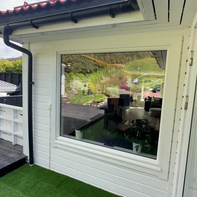 Hvit boligfasade med stort vindu, takrenne og overbygd terrasse med grønt kunstgress.
