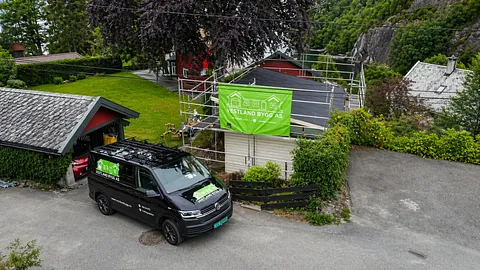 Firma­bil parkert ved bolig under rehabilitering, med stillas og profilert byggskilt på fasaden.