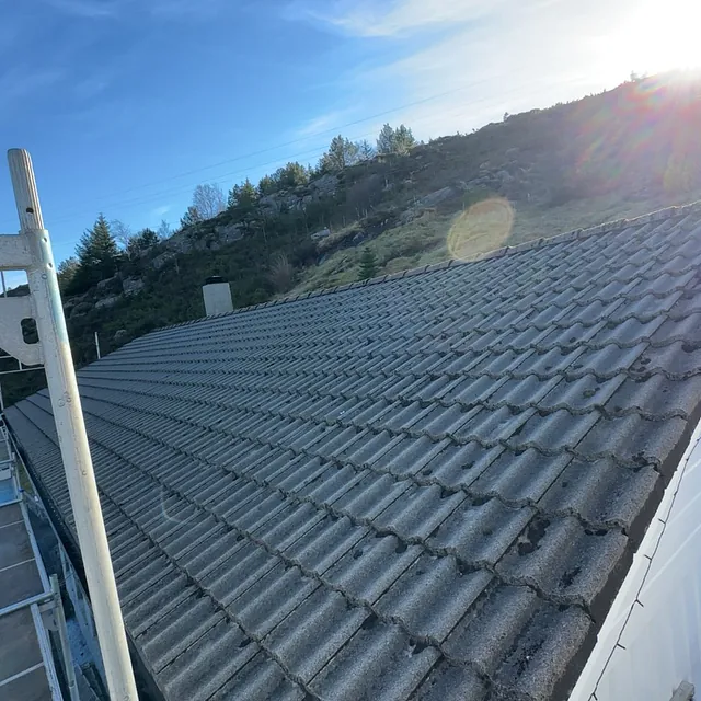 Mørkt tak med taksteinprofil fotografert fra stillas i sollys, med åsside og blå himmel i bakgrunnen.