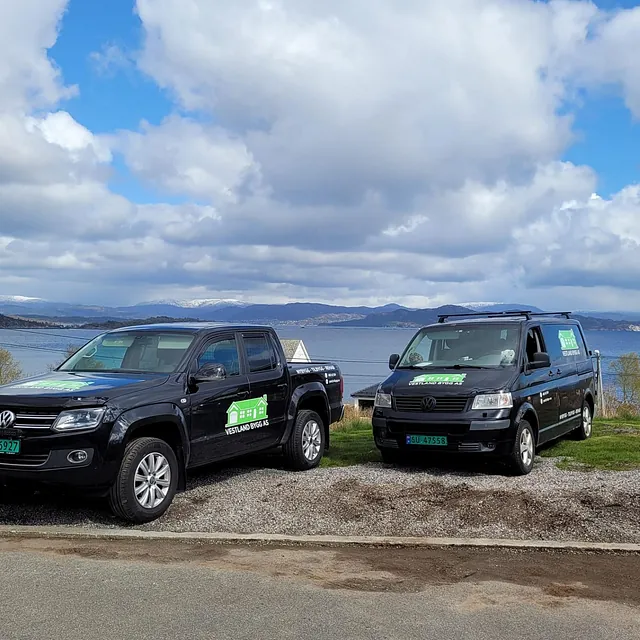 To profilerte firmabiler parkert ved sjøen med fjellutsikt og delvis skyet himmel i bakgrunnen.